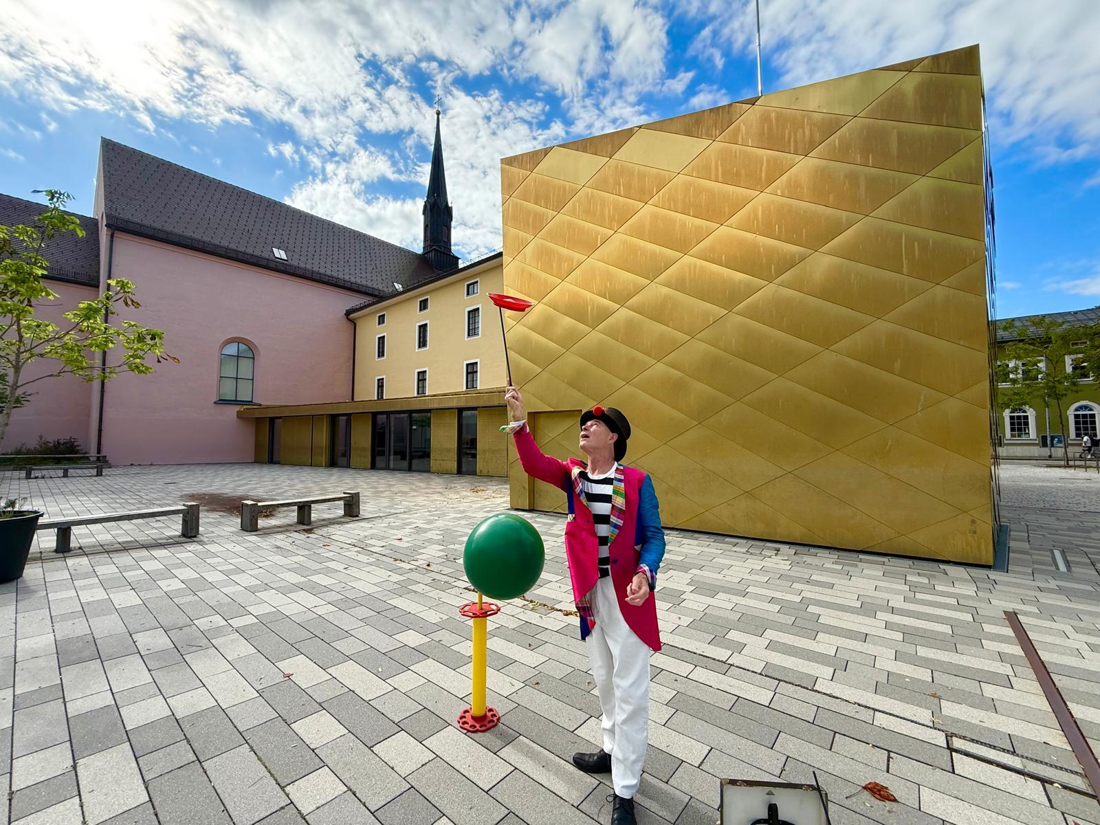A clown in a colorful suit spins a plate on a stick in a modern plaza between striking buildings.