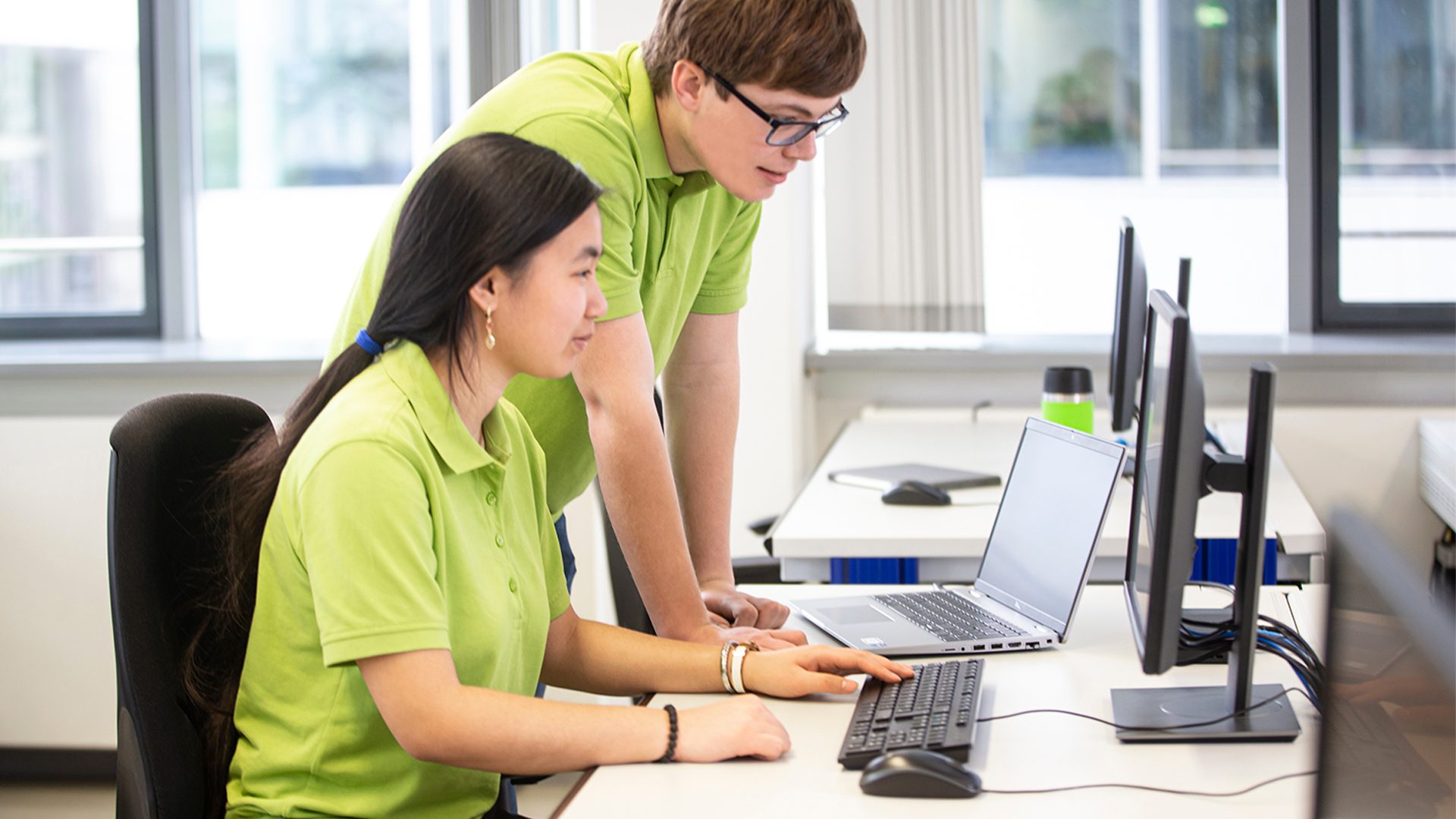 Two young people in green shirts collaborate on a laptop in an office.