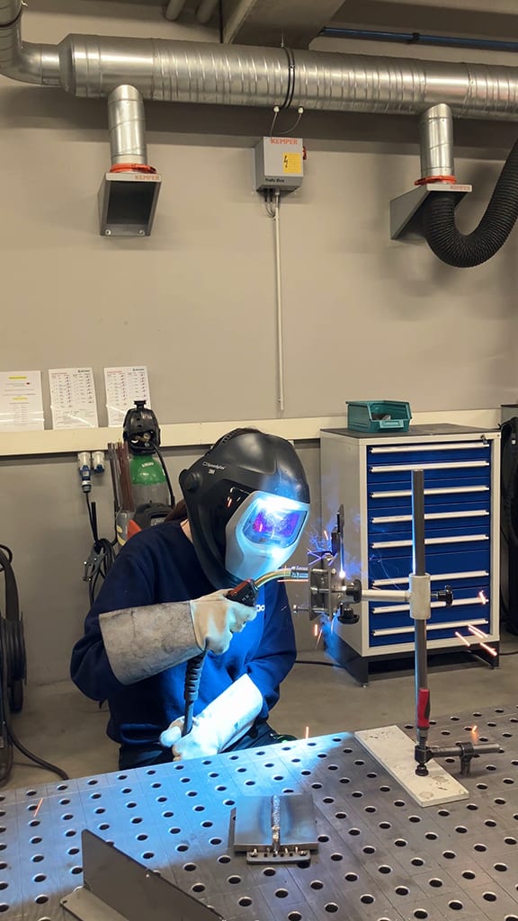 Person welding in a workshop, wearing a helmet and gloves, with sparks flying.