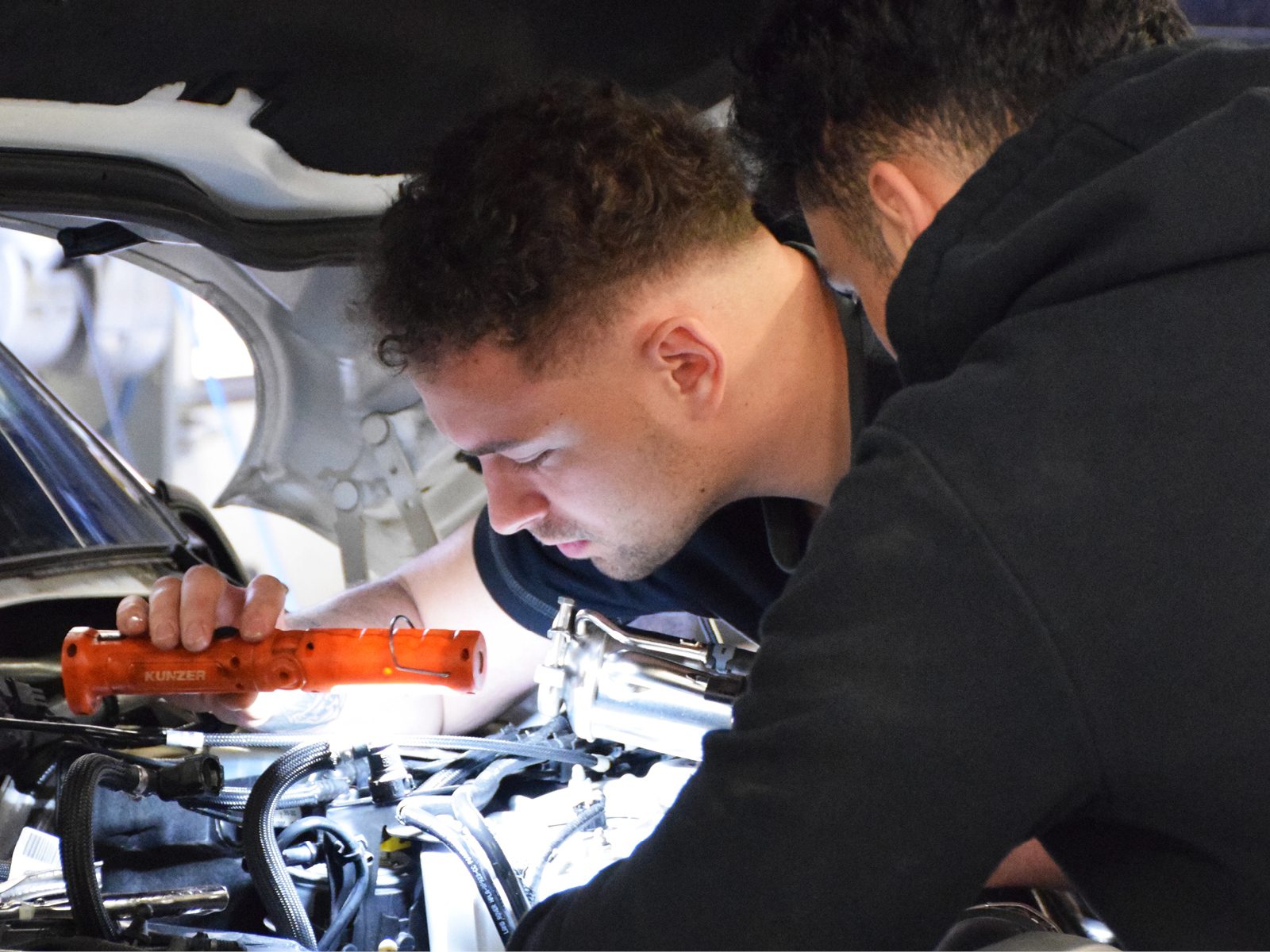 Two mechanics inspecting a car engine, one holding a flashlight.