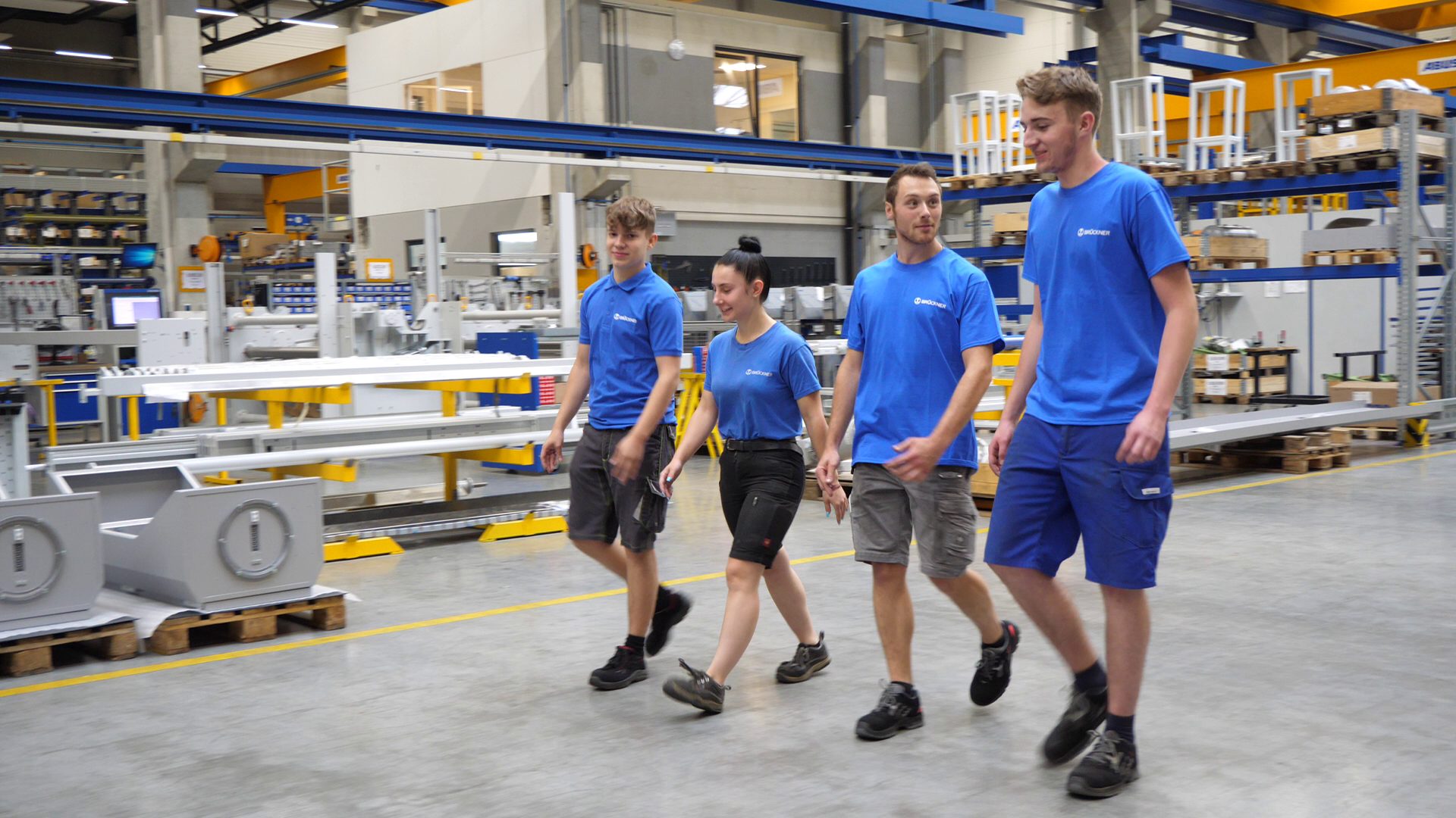Four young factory workers in blue shirts walking through a manufacturing plant.