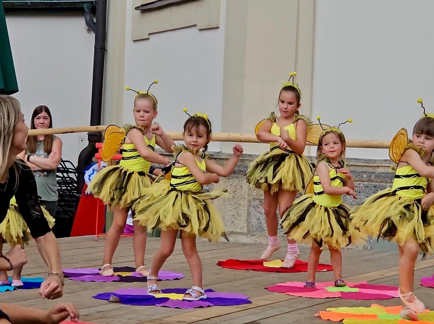 Young girls in bumblebee costumes perform a dance on a wooden stage, with an adult supervising.