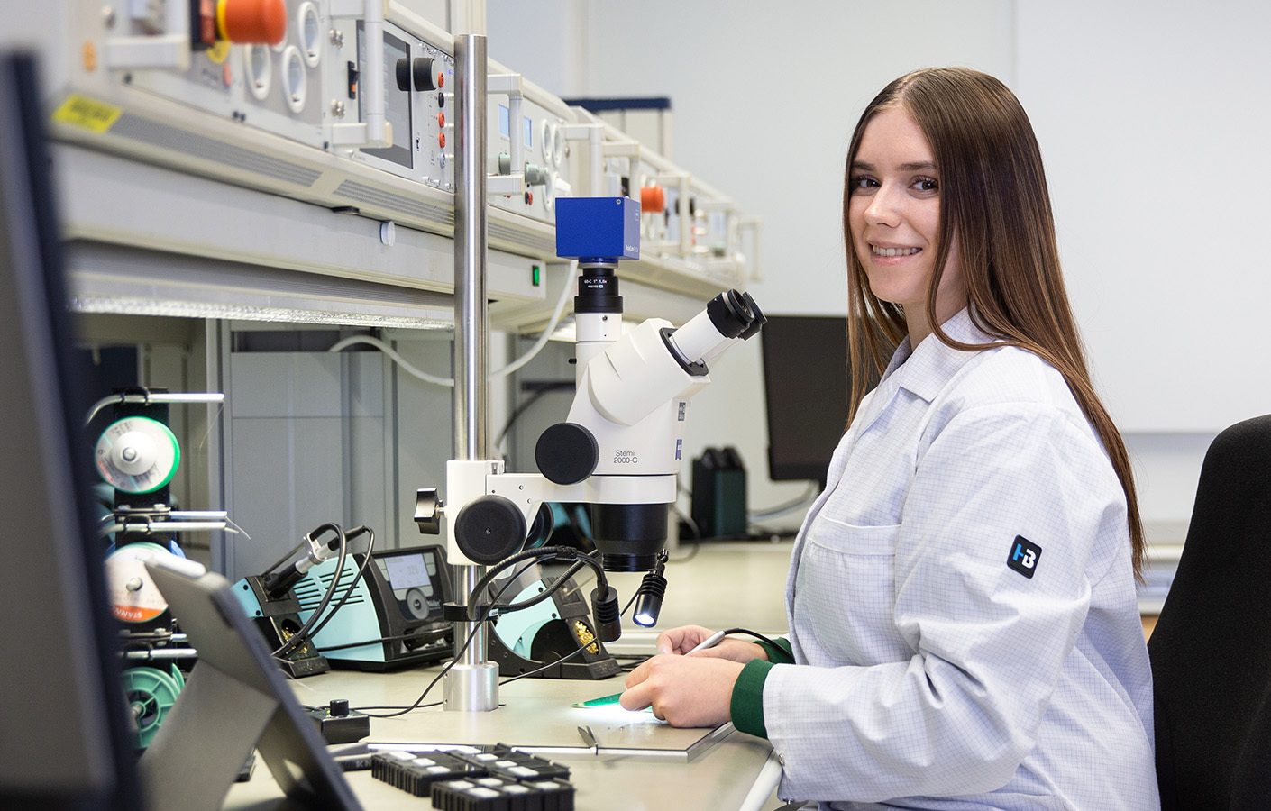 Smiling woman in a lab coat working with a microscope and electronics.