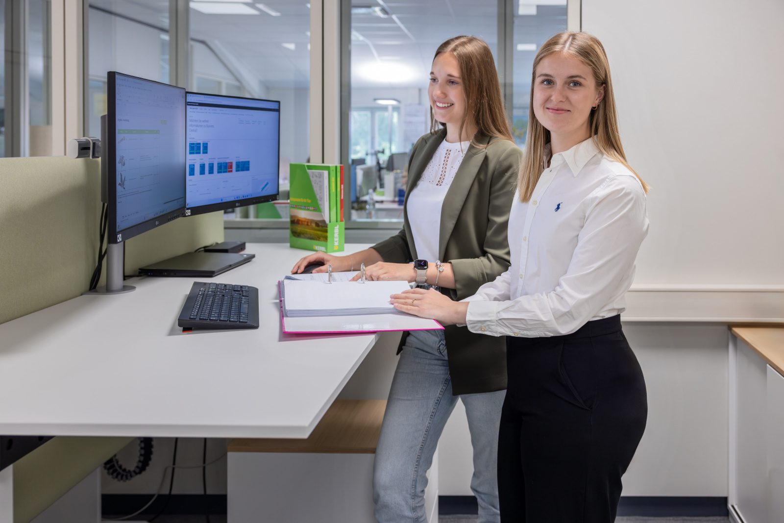 Two young women stand smiling at a modern office desk with dual monitors and binders.