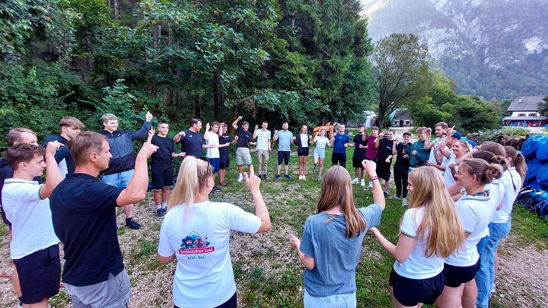 A group of people stand in a circle outdoors, many with arms raised or pointing, playing a game.