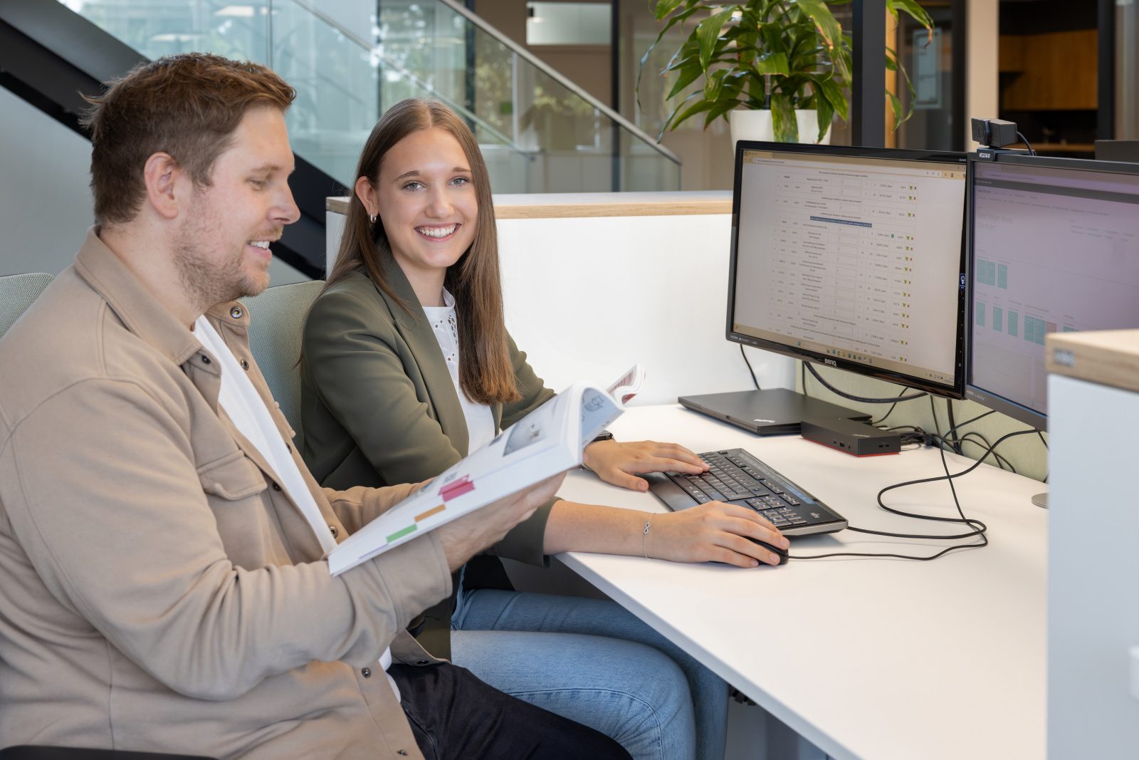 Two colleagues at an office desk with computers, one smiling at the camera while the other holds a document.