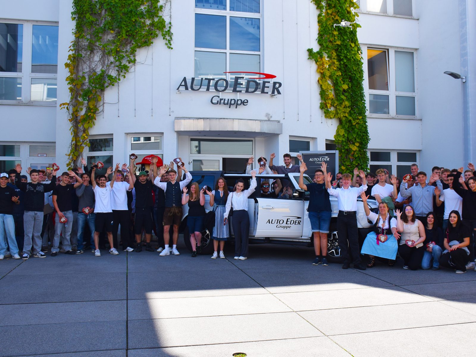 A large group of young people waves in front of the AUTO EDER Gruppe building and a white car.