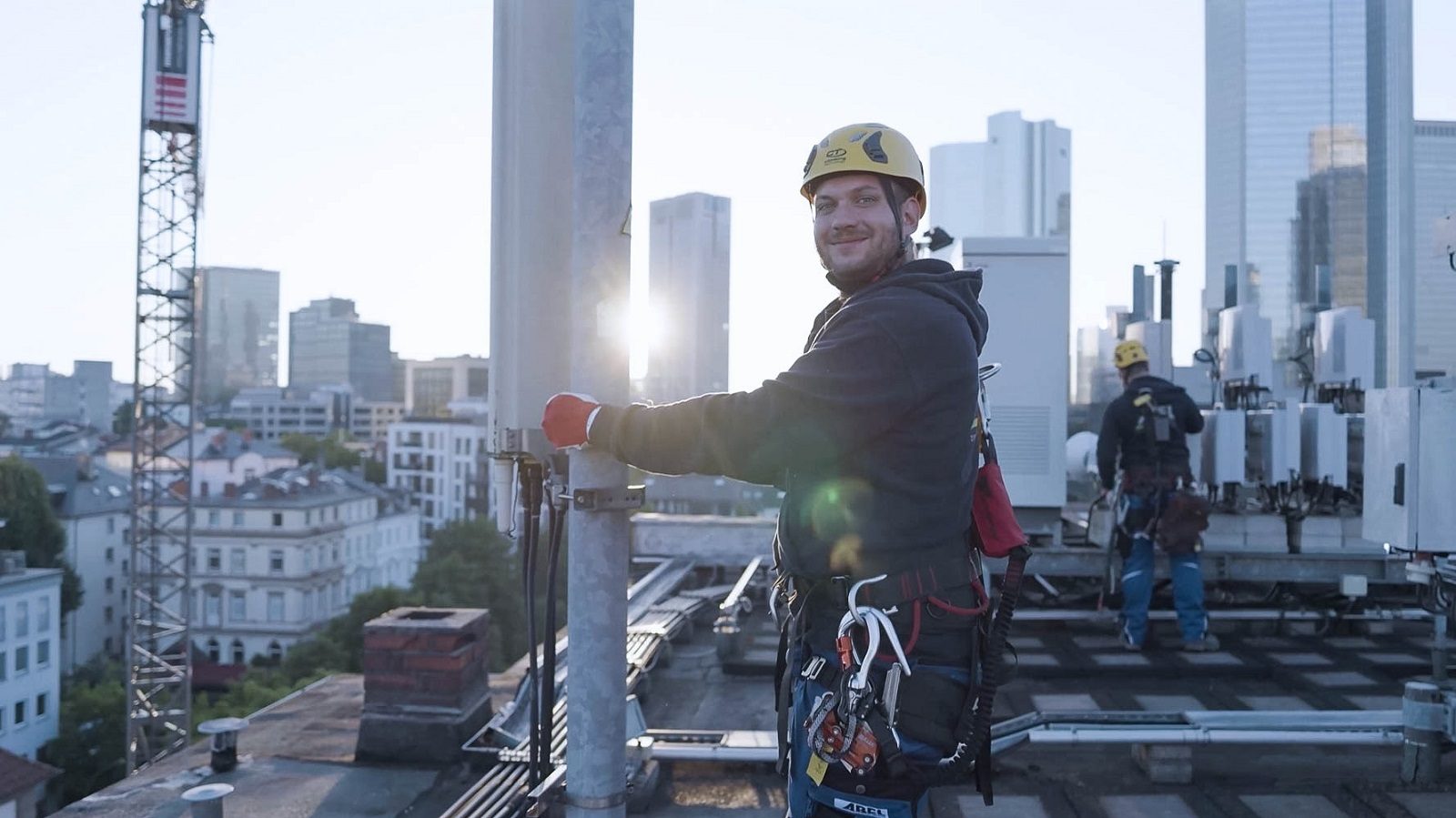 Smiling worker in safety gear on a city rooftop with buildings and telecom equipment.