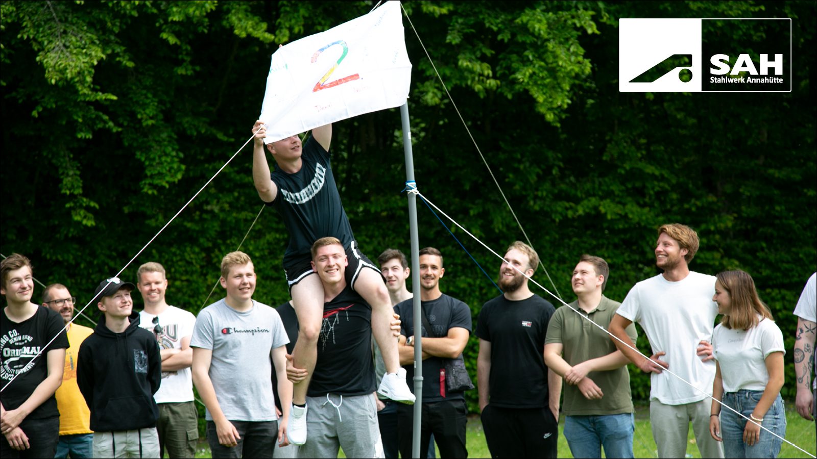 A group of young people outdoors; one on another's shoulders holds a flag with the number '2'.