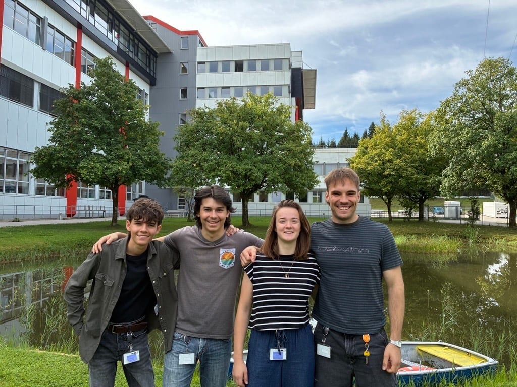 Four smiling students, two men and two women, pose outdoors in front of a school building and pond.