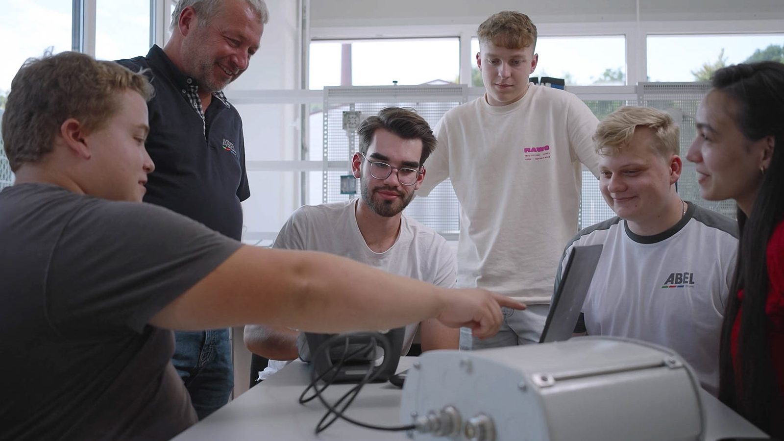 Six people, young and older, collaborating around a table, pointing at a technical device.