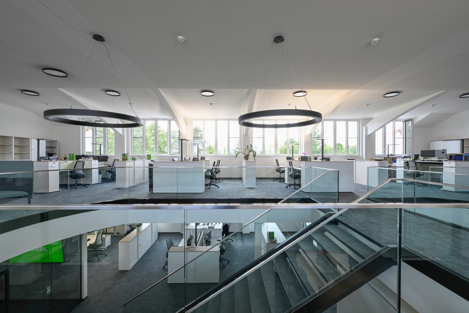 Modern two-story open-plan office with desks, large windows, glass stairs, and circular lights.