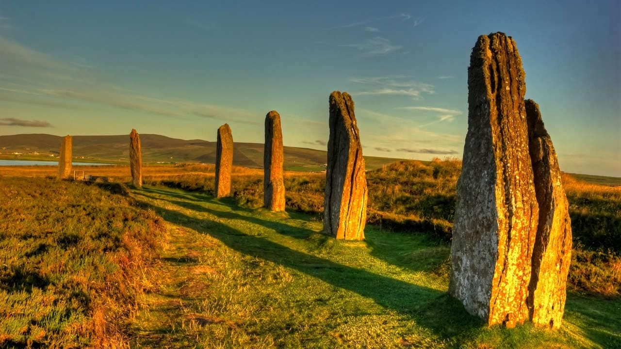 Ancient standing stones in a grassy field at sunset, with long shadows and a distant lake under a clear sky.
