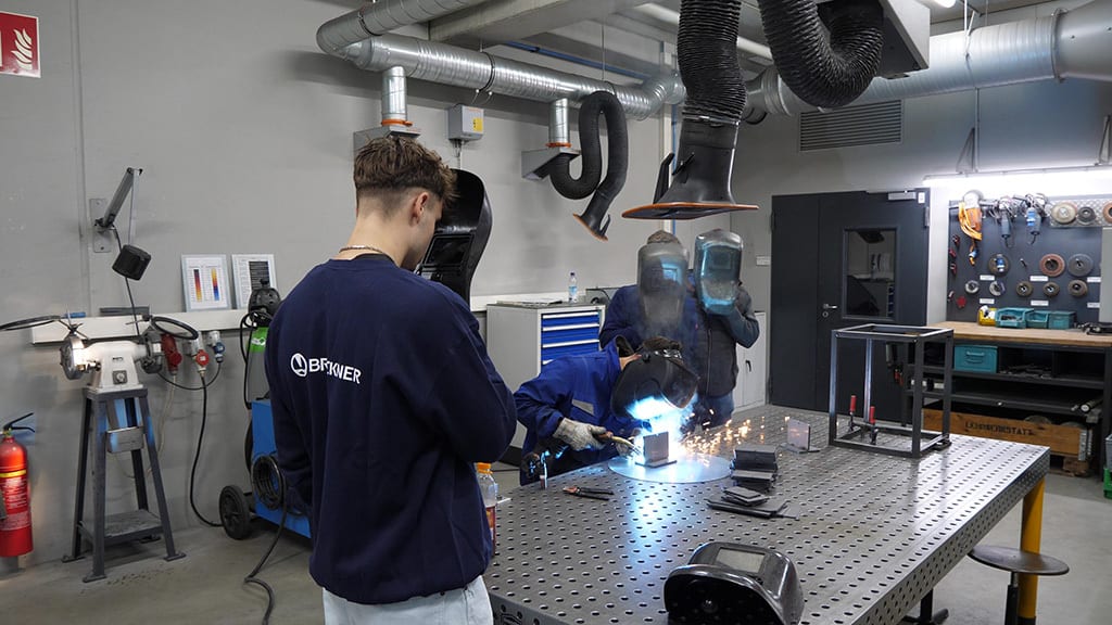 People welding and observing in a busy workshop, sparks flying from active welding.