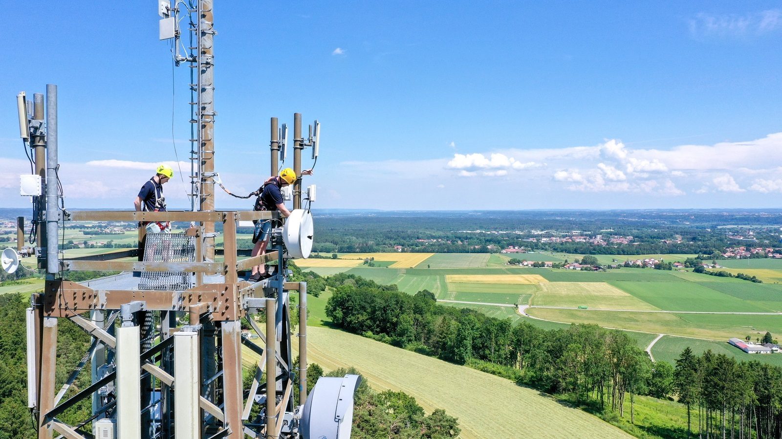 Two technicians maintain a cell tower, with a panoramic view of fields and distant towns below.