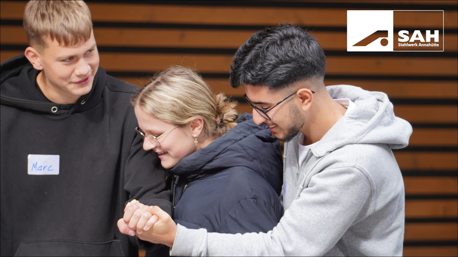 Three young people, including Marc, interact and look down, SAH logo in corner.