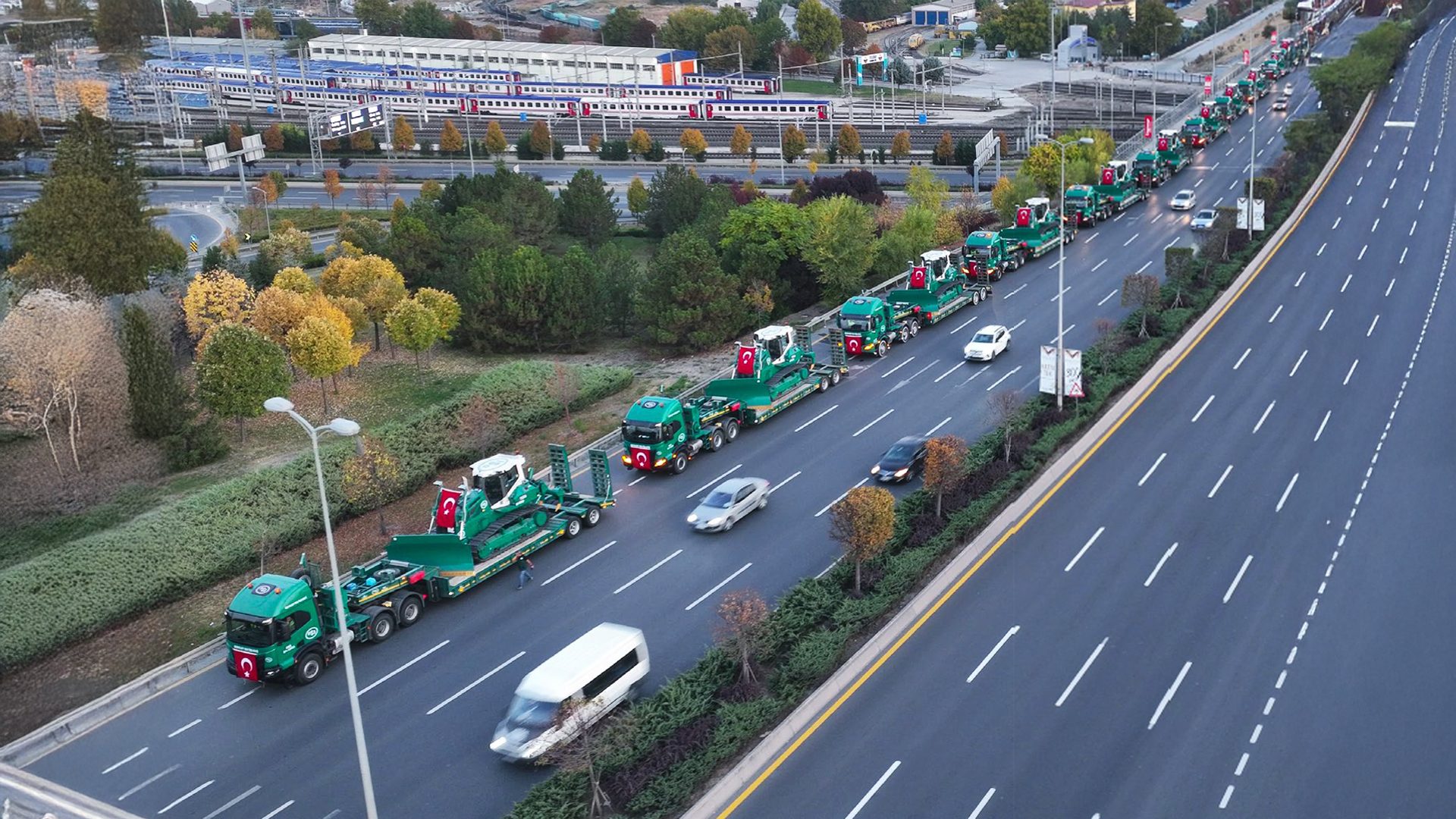Long convoy of green trucks with Turkish flags carrying construction equipment on a highway.