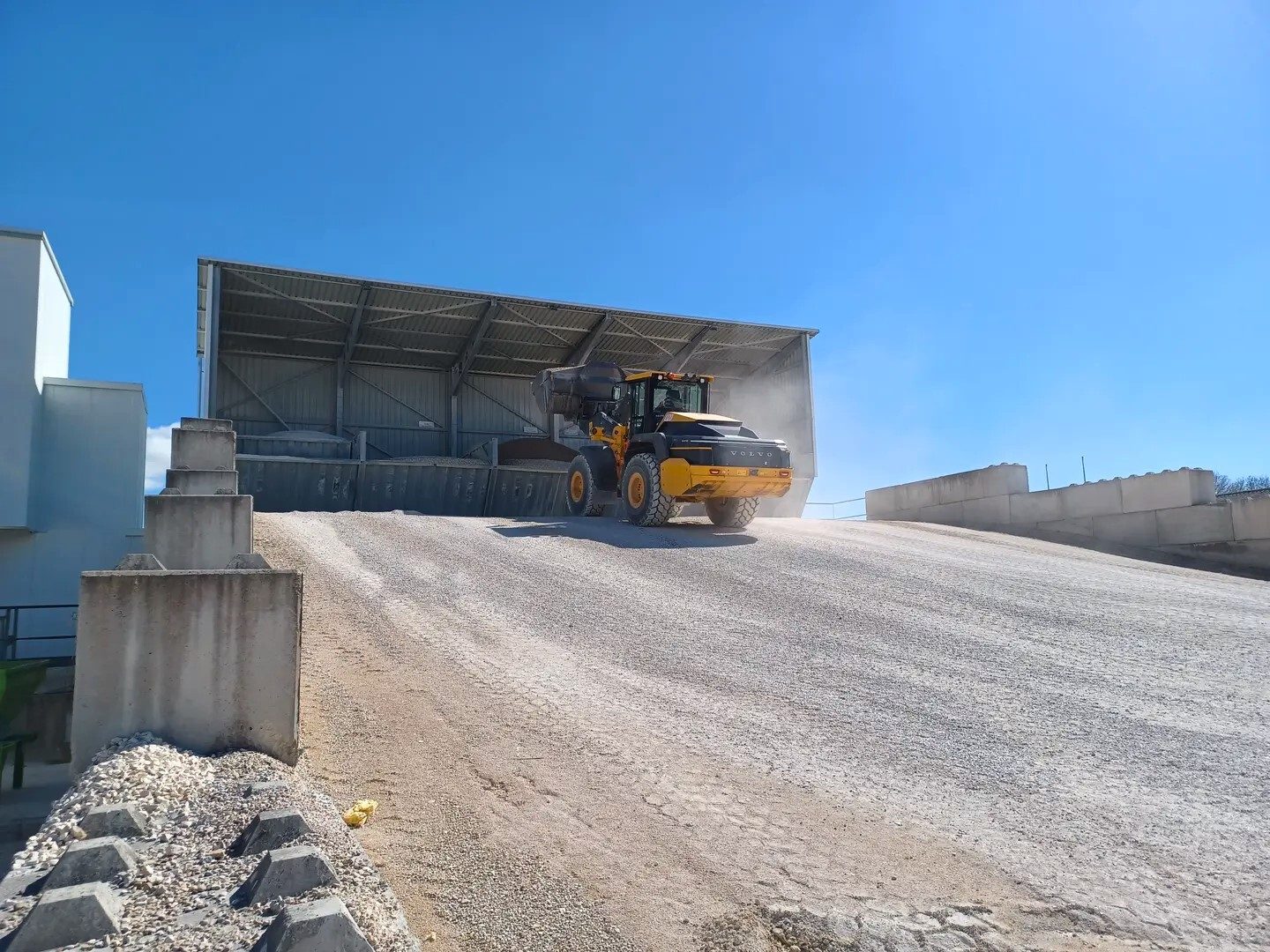 A Volvo front loader drives up a dusty gravel ramp towards a large storage shed under a blue sky.