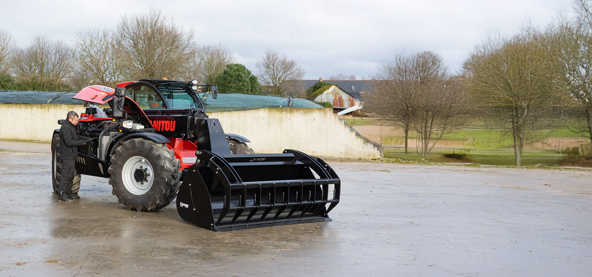 Man inspects a red and black Manitou telehandler with a large bucket attachment in a farmyard.