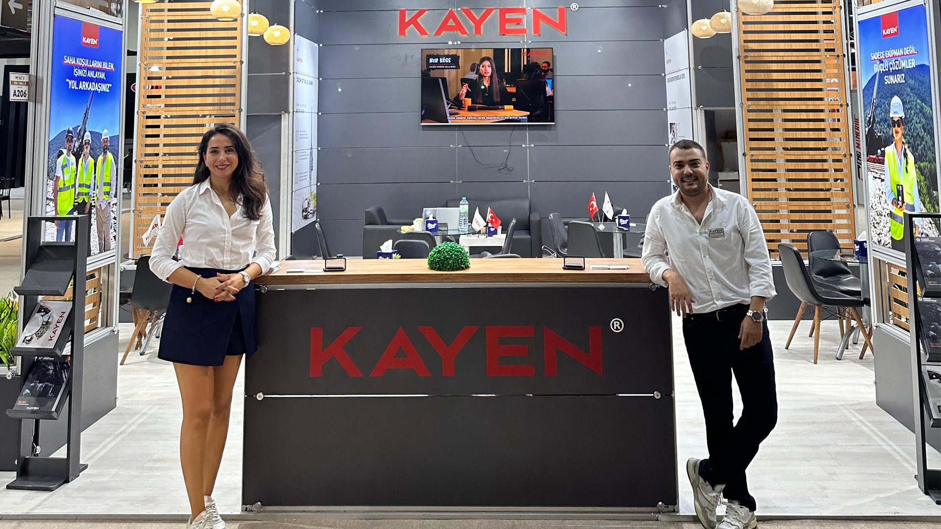 Two smiling employees, a man and a woman, stand at a KAYEN booth, showcasing products and services.