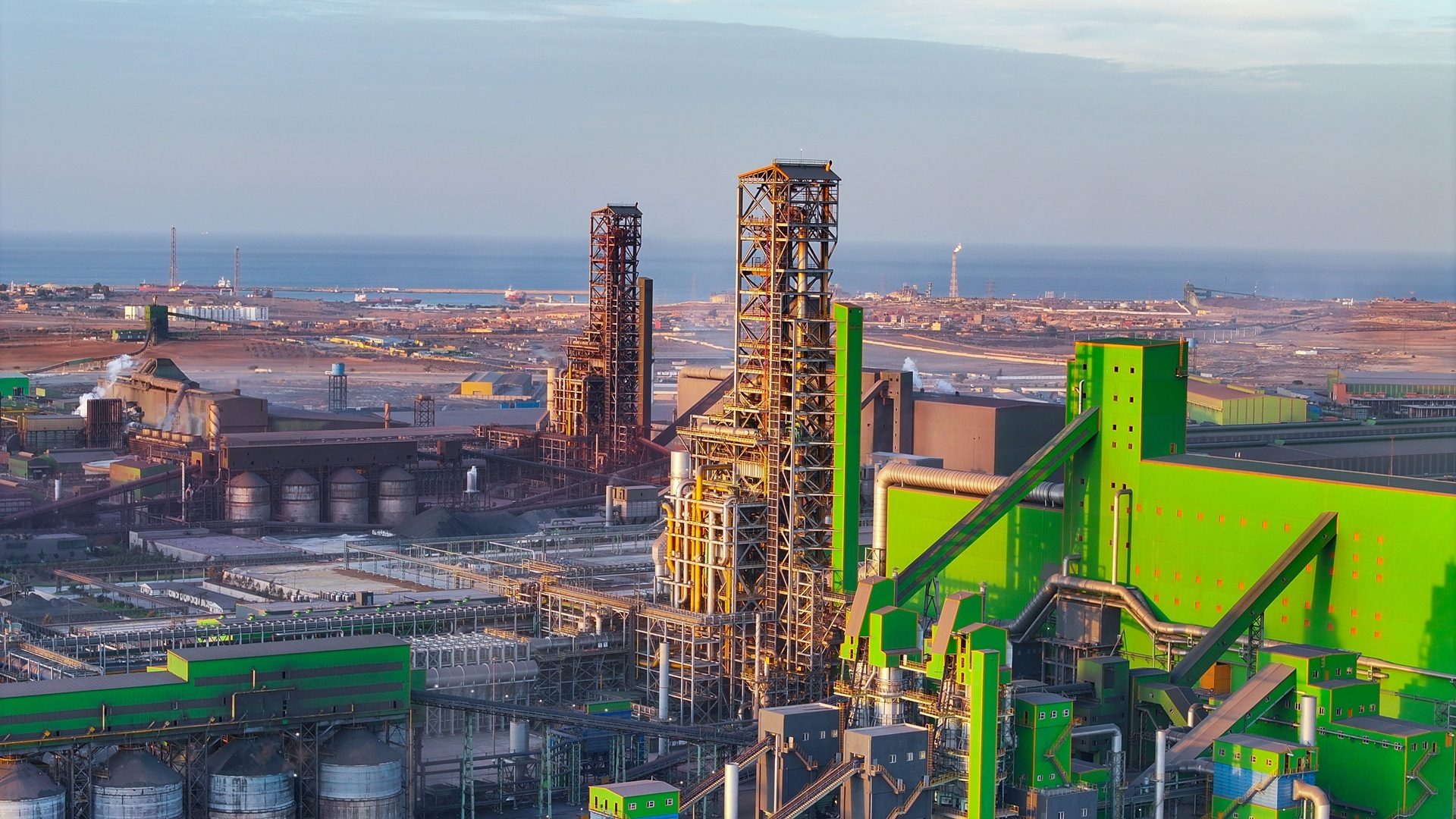 An aerial view of a large industrial complex with tall structures and green buildings, located near the sea.