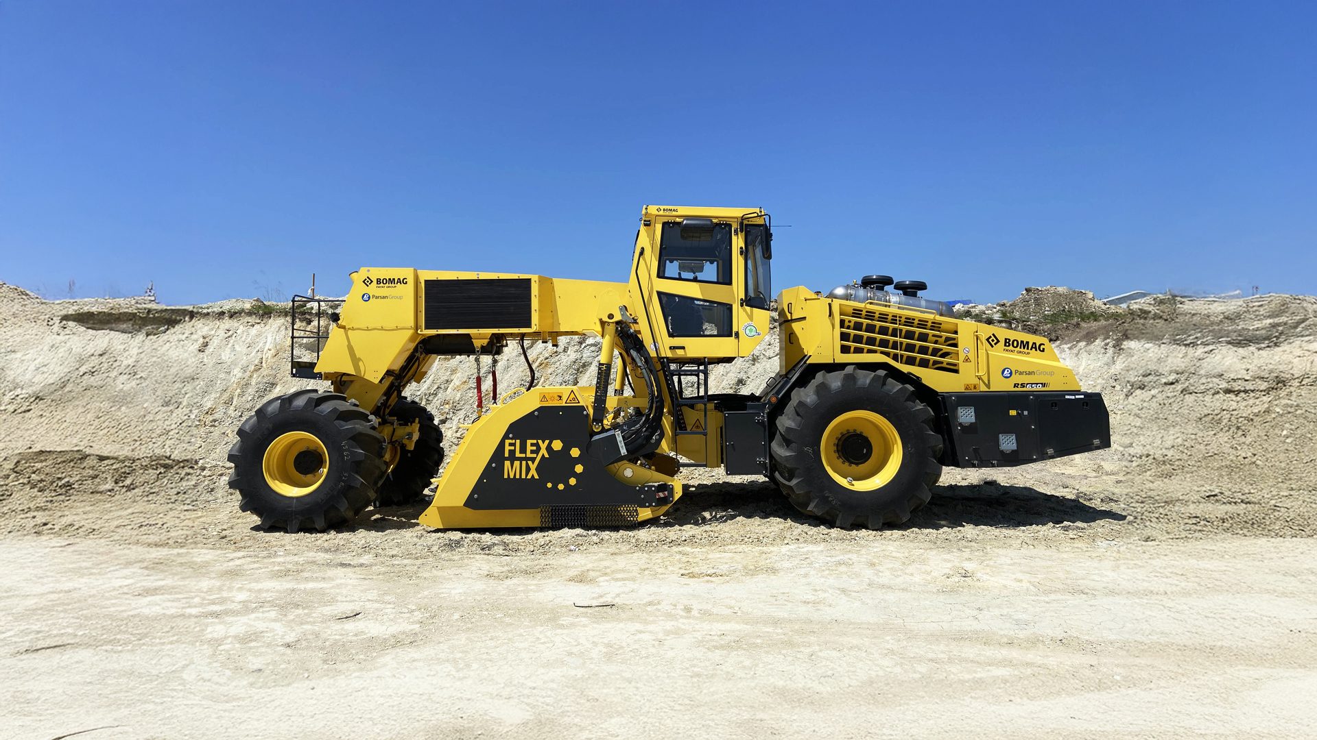 Yellow BOMAG Flex Mix construction machine on a sandy site under a clear blue sky.