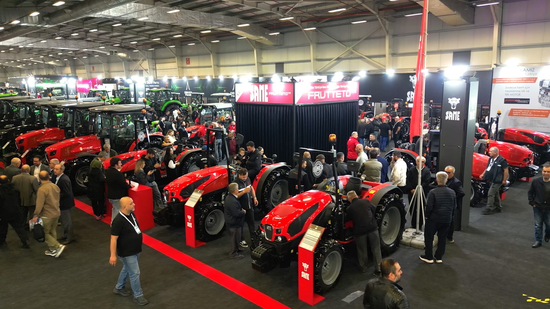 Crowded indoor exhibition hall with red and green tractors on display, featuring brands like SAME.