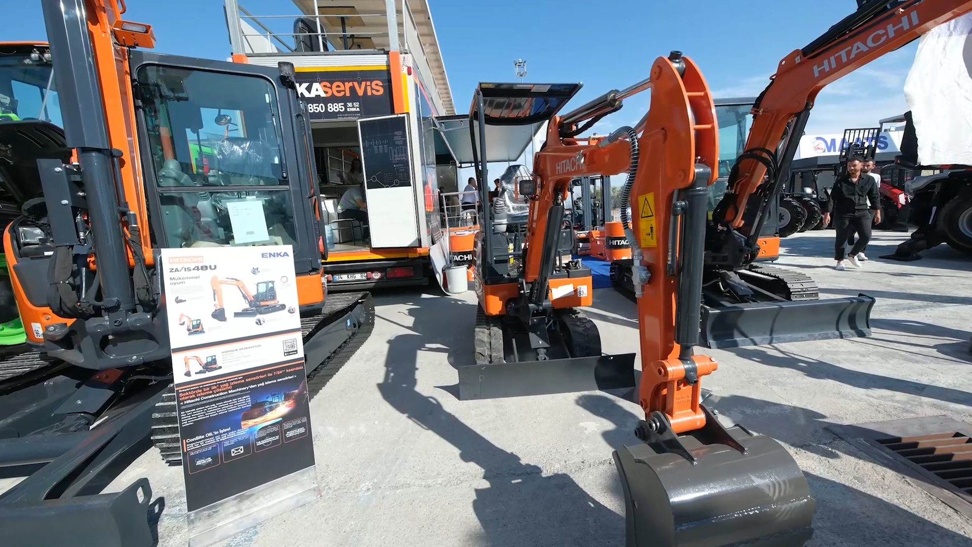 Orange Hitachi excavators on display with a branded truck and information stand.