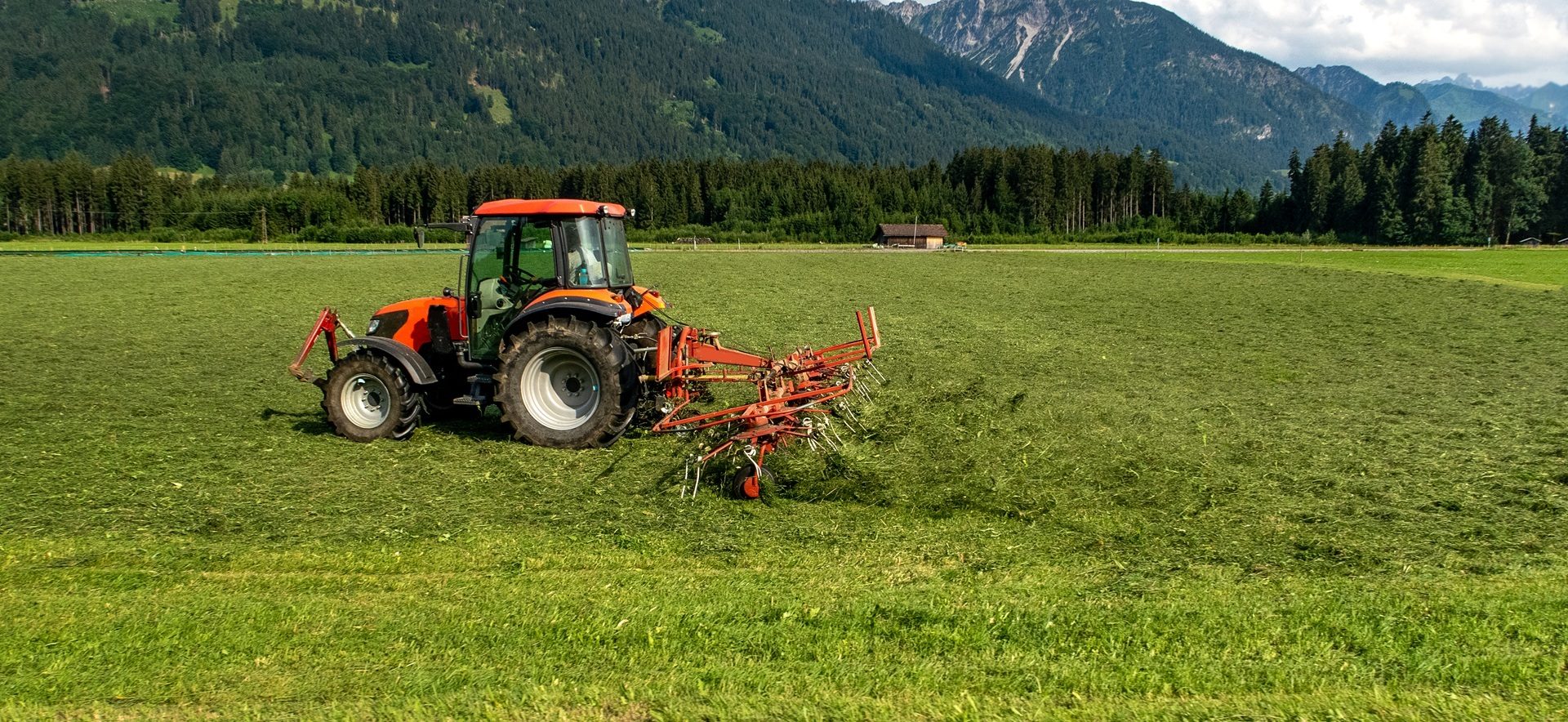 Orange tractor raking hay in a field, with mountains and trees in the background.