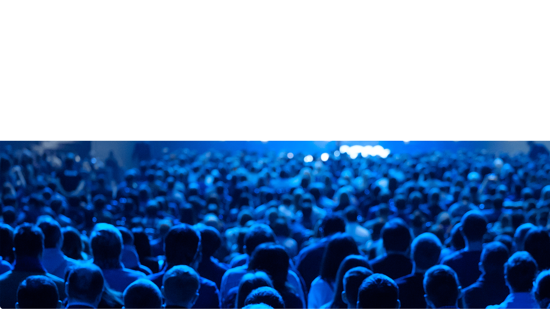 Audience at an event, seen from behind, under blue stage lights.