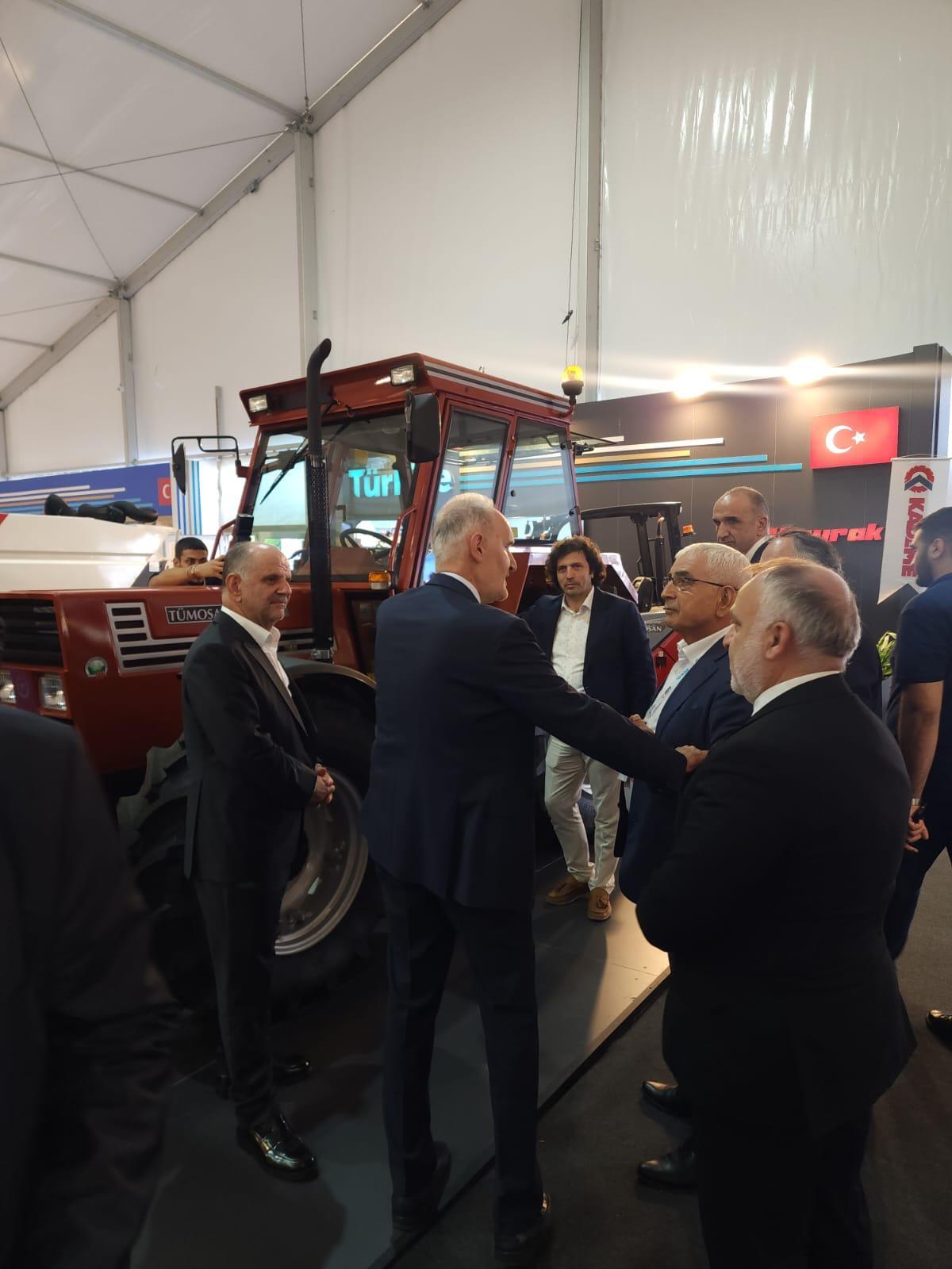 Men in suits converse by a red TÜMOSAN tractor at an indoor exhibition.