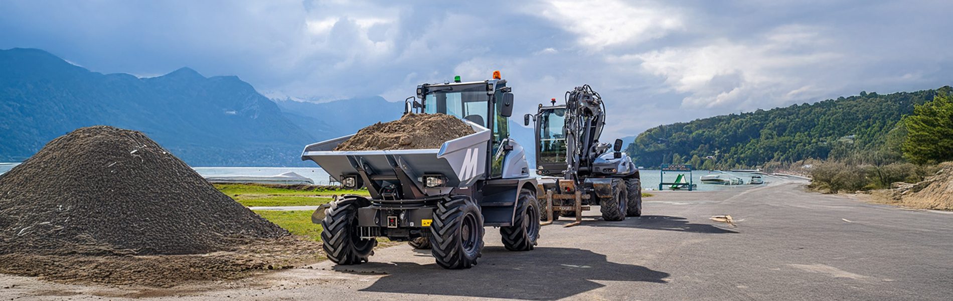 Two construction vehicles, a dumper and an excavator, on a road beside a lake with mountains.