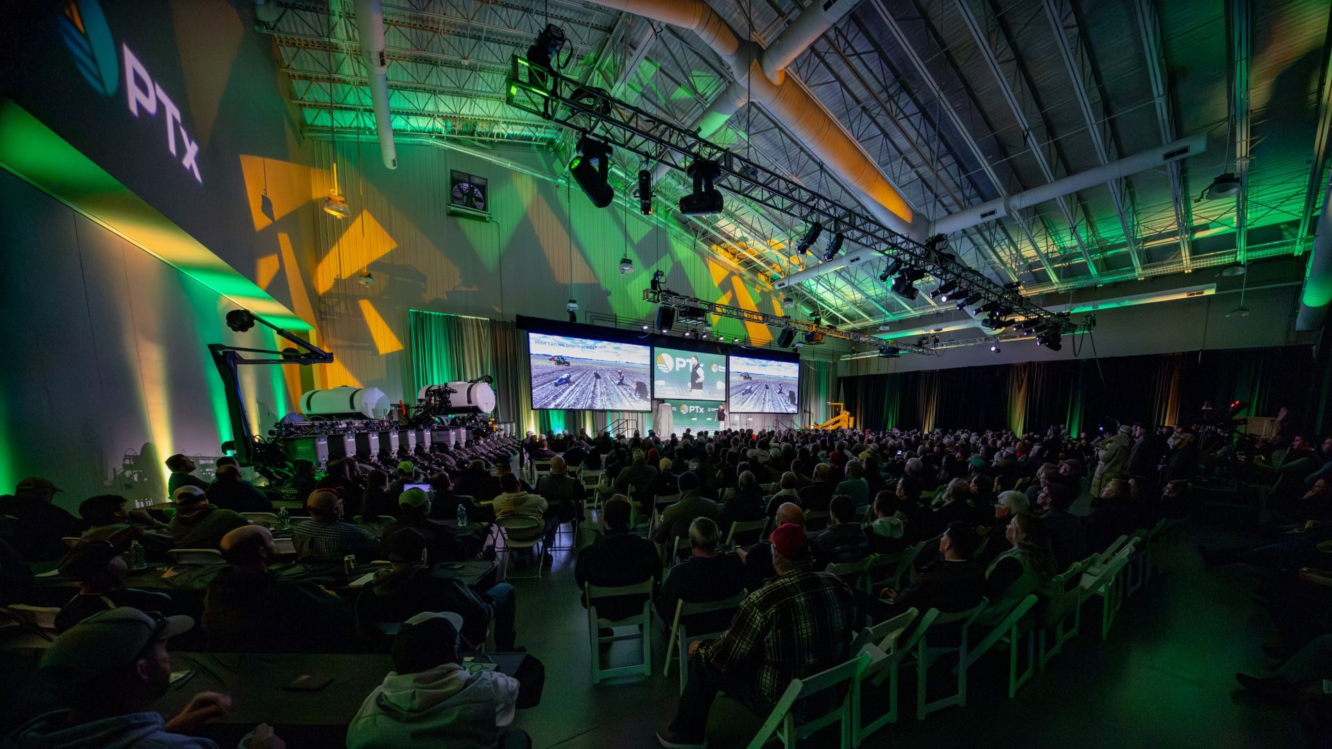 Large agricultural conference: audience, PTx screens, farm machinery on stage under green and yellow lights.