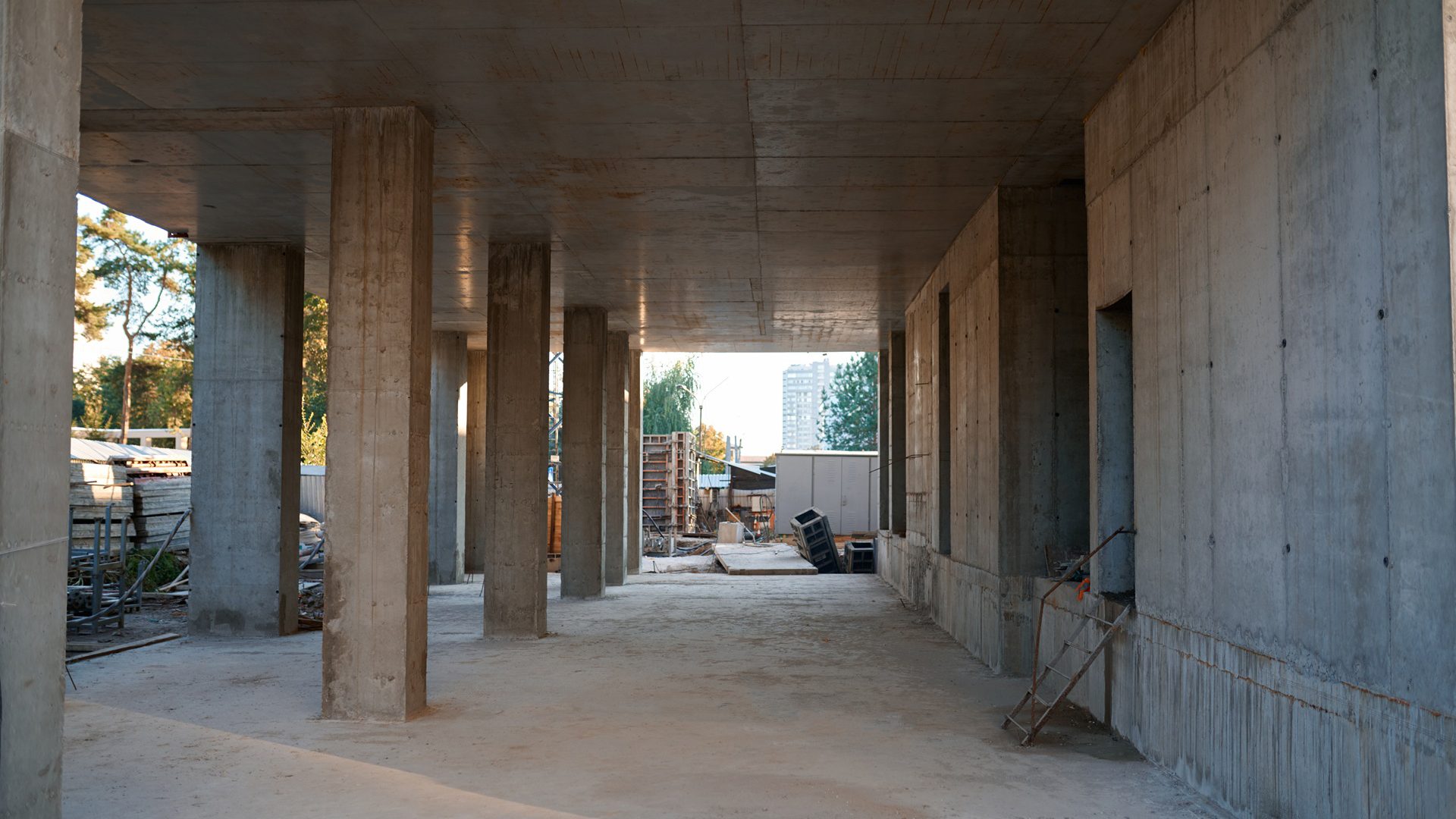 Unfinished concrete building with pillars, a long wall, and an open view to trees and another building.