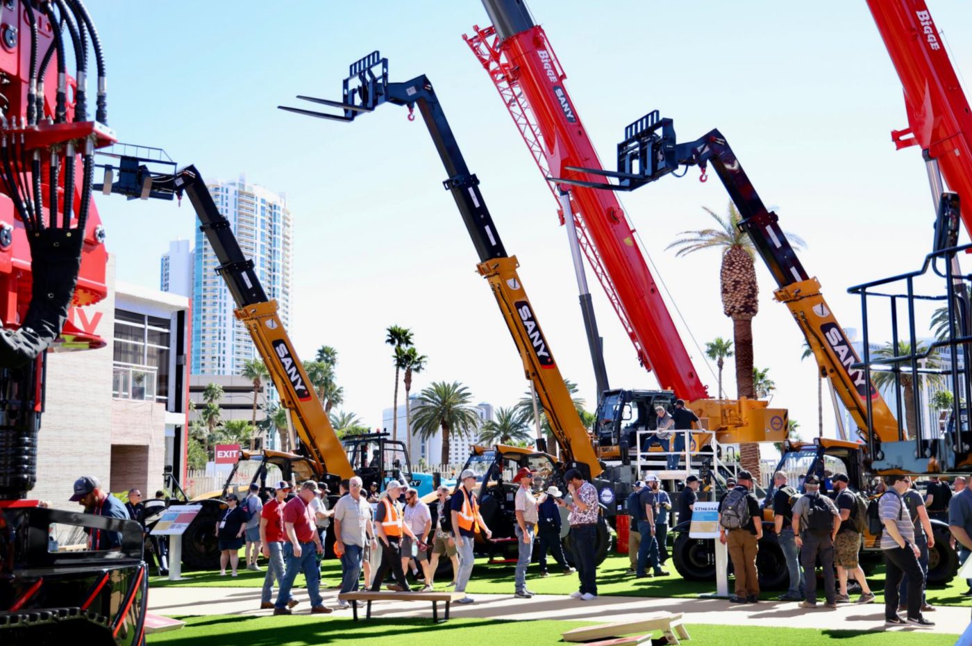 People view SANY telehandlers and cranes at an outdoor exhibition, with city buildings and palm trees.