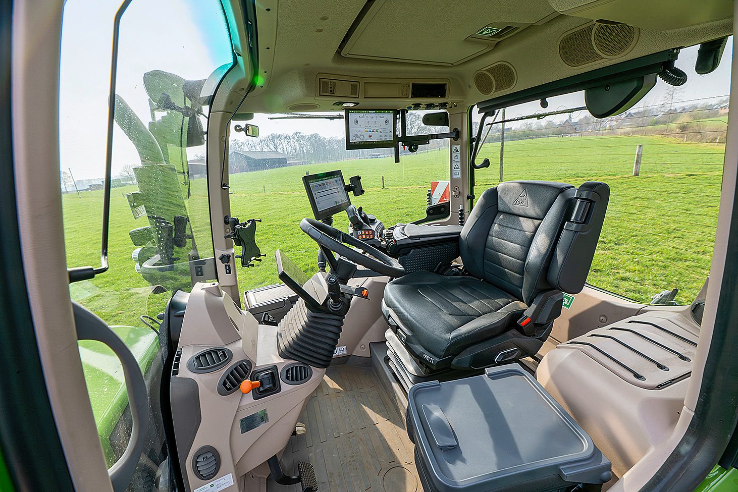 Modern tractor cabin interior with driver's seat, multiple screens, controls, and green fields outside.