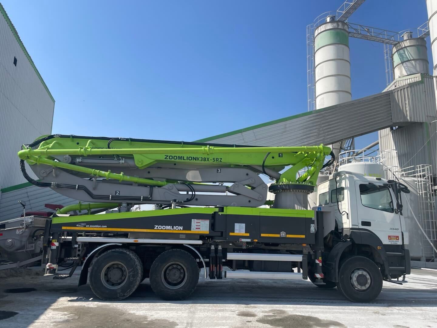 A green and white Zoomlion concrete pump truck parked at a concrete batching plant under a clear blue sky.