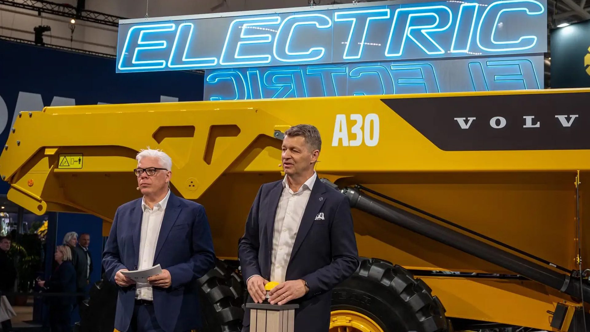 Two men present a yellow Volvo A30 electric hauler with an 'ELECTRIC' neon sign in the background.