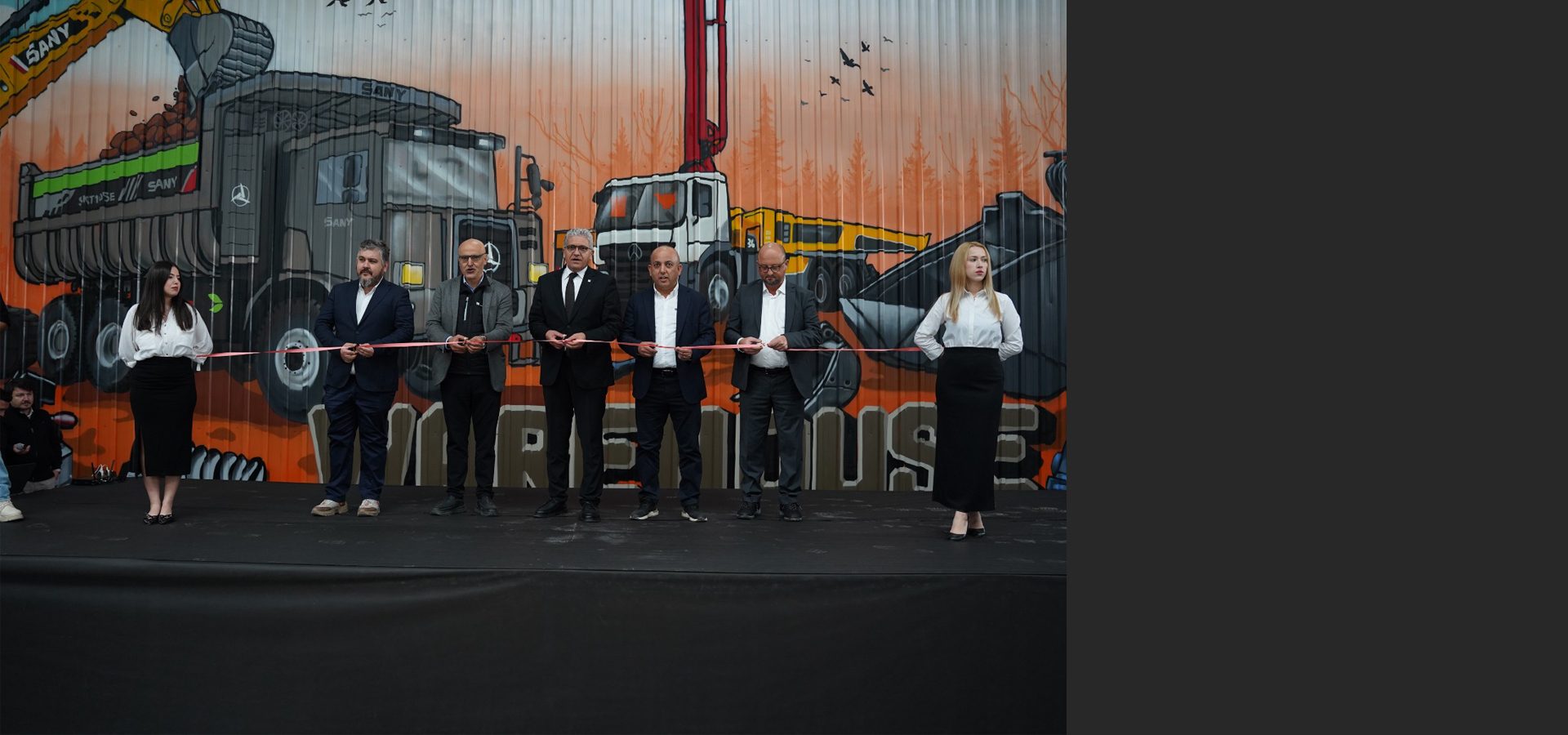 A group of people, mostly men, cutting a pink ribbon in front of a colorful mural of construction vehicles.