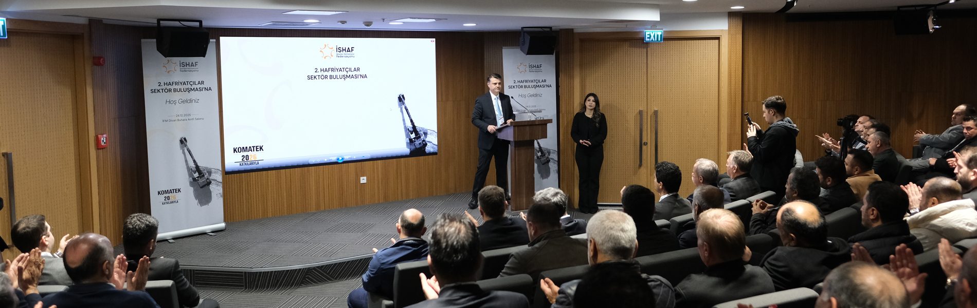 Speaker presents at a business conference with İSHAF and KOMATEK logos on screen, audience applauds.