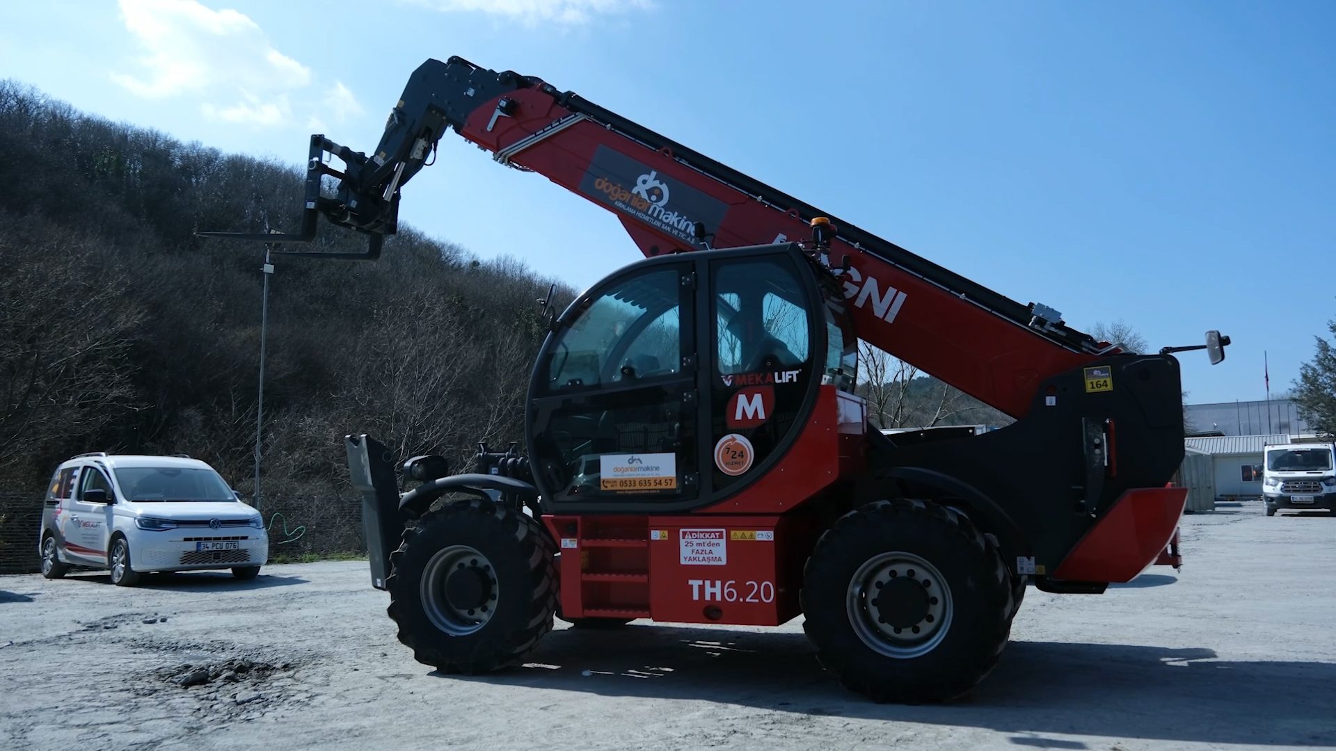 Red Magni telehandler with an extended boom and forks, parked on a gravel lot next to two white vans.