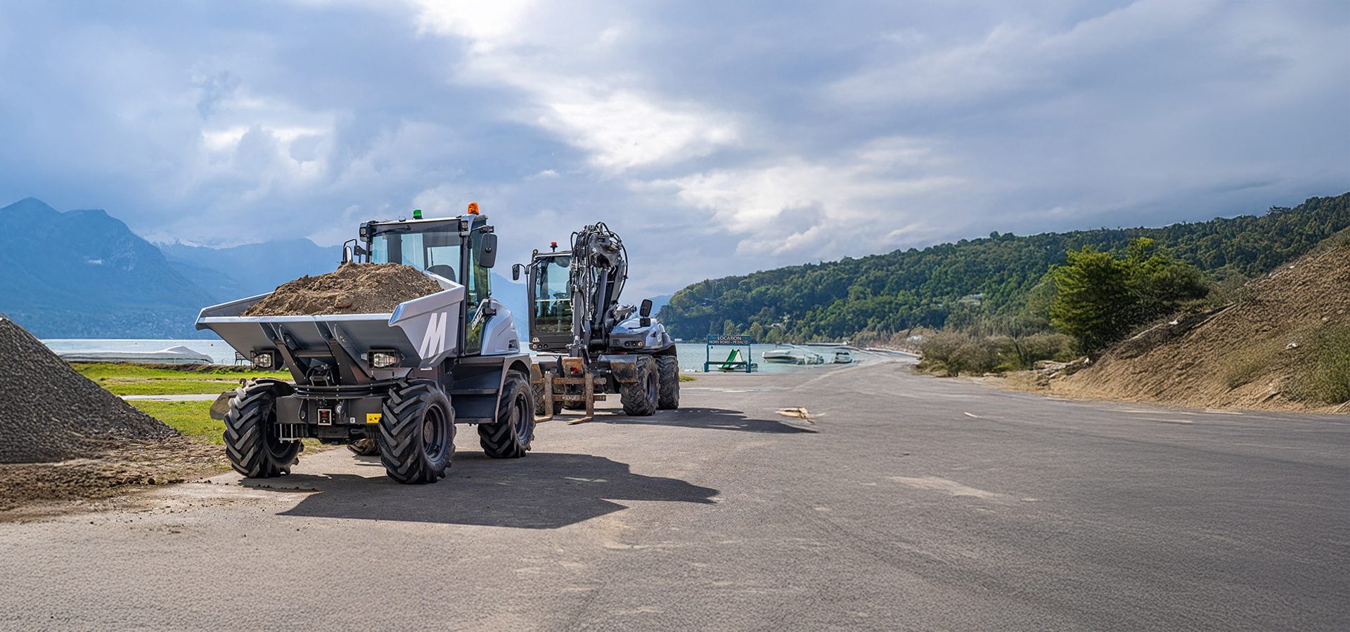 Dumper with dirt and excavator by a lake with mountains under a cloudy sky.