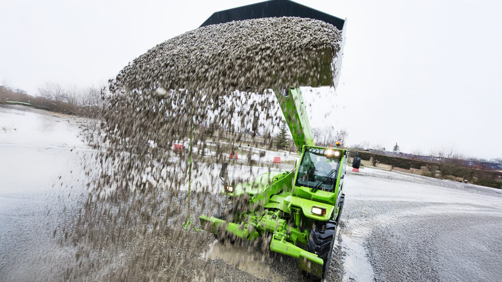 Bright green telehandler dumping gravel in a blurry cascade onto wet, grey ground.