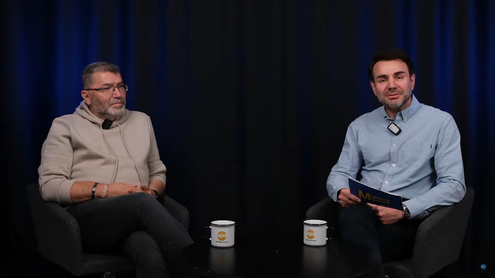 Two men sit in a studio with a dark blue background, both wearing microphones.