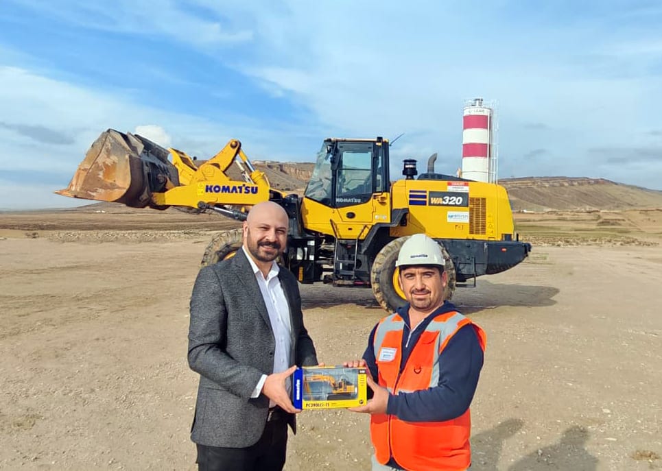 Two men, one in a suit and one in a hard hat, present a Komatsu model in front of a Komatsu WA320 loader.