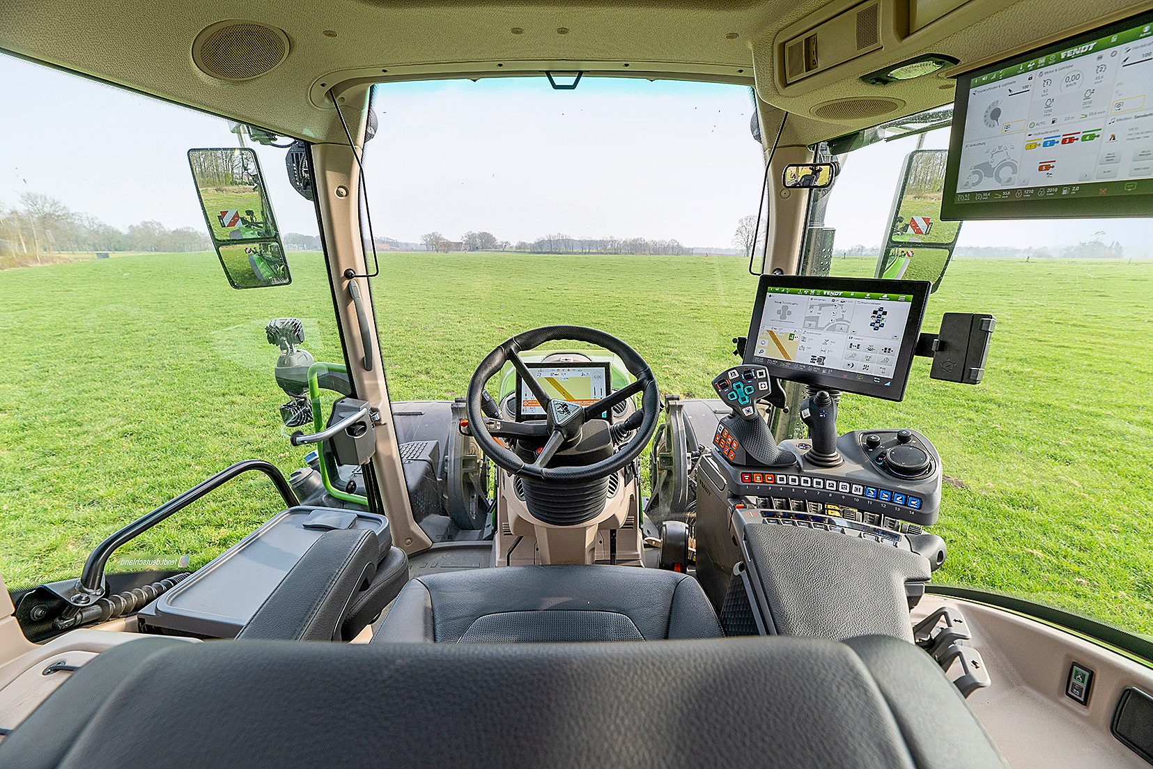 View from inside a modern Fendt tractor cab, showing multiple digital screens, controls, and a green field outside.