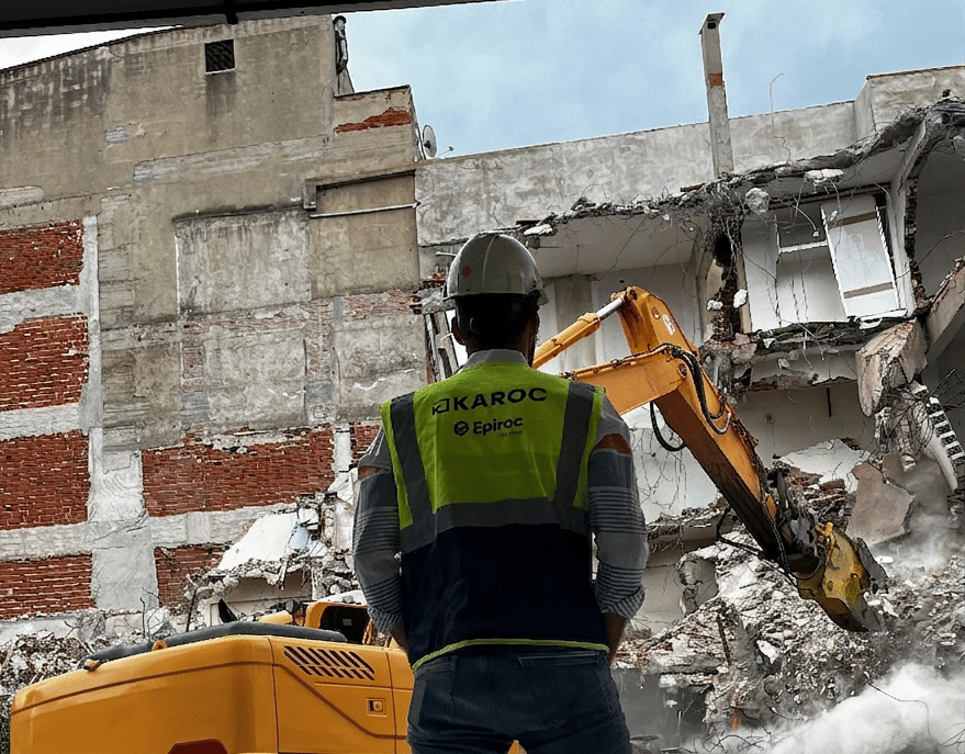 Worker watches excavator demolish a building, rubble and dust visible.