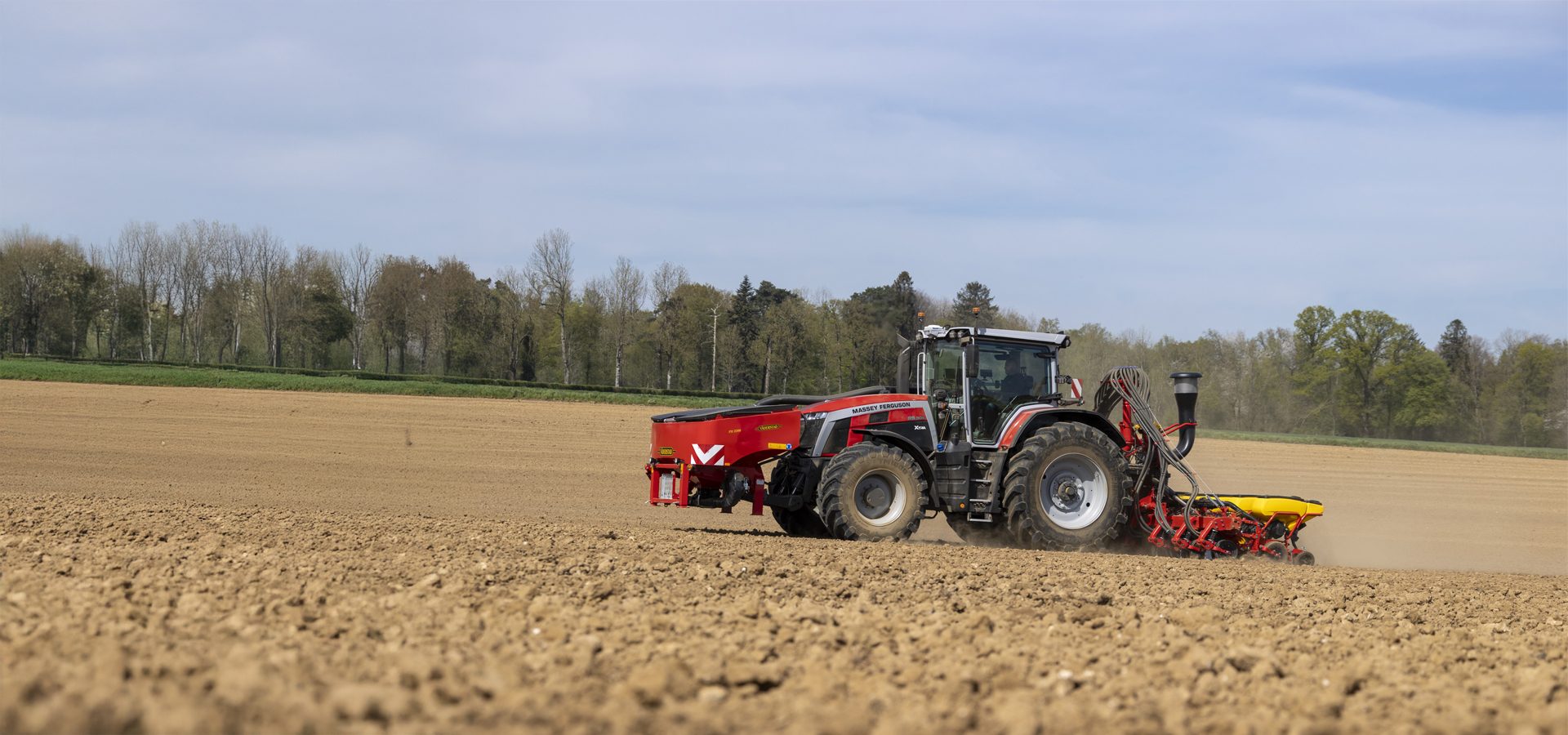 A red and grey Massey Ferguson tractor pulls a seeder across a brown, freshly tilled field under a blue sky.