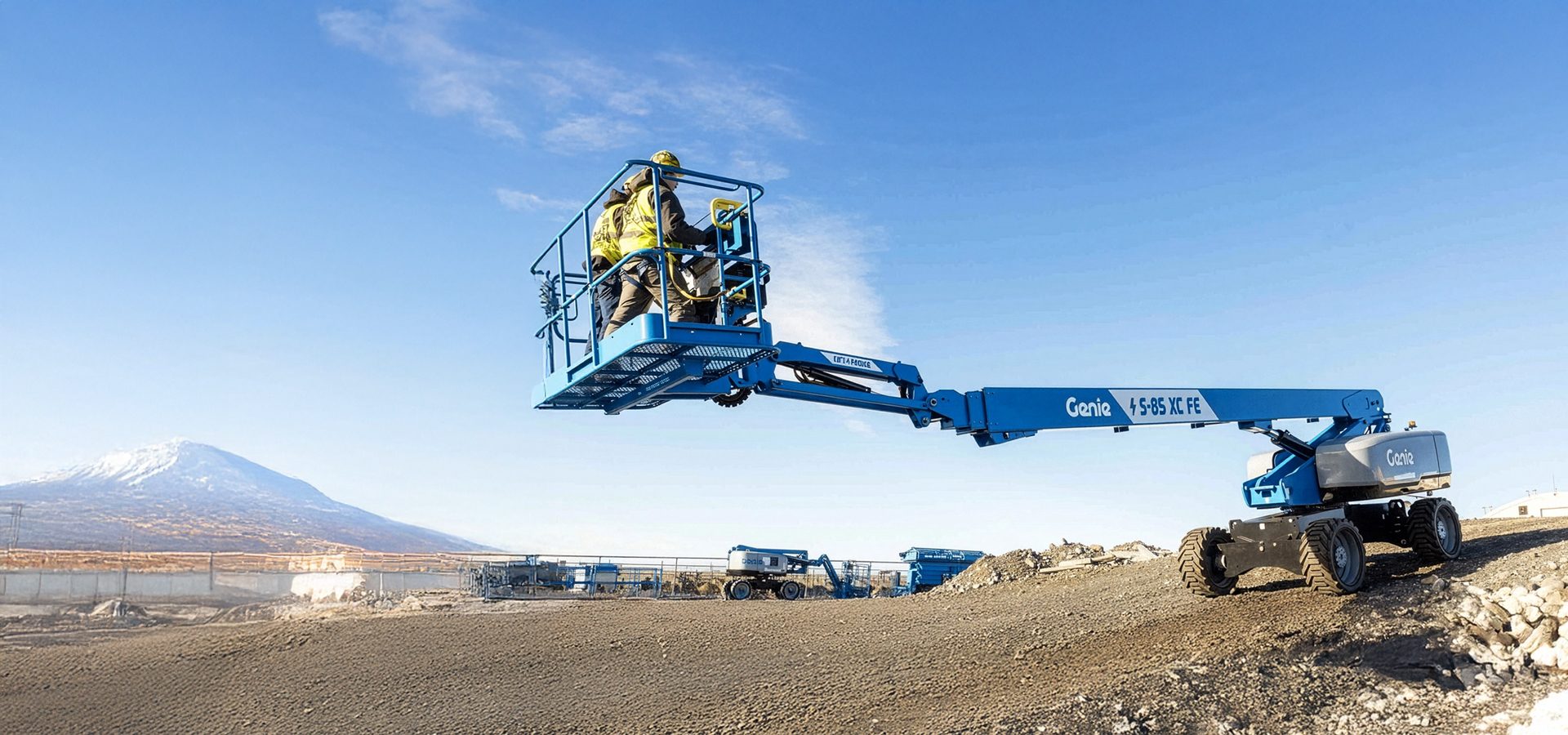 Workers operate a blue Genie boom lift at a construction site under a blue sky, with a snowy mountain.