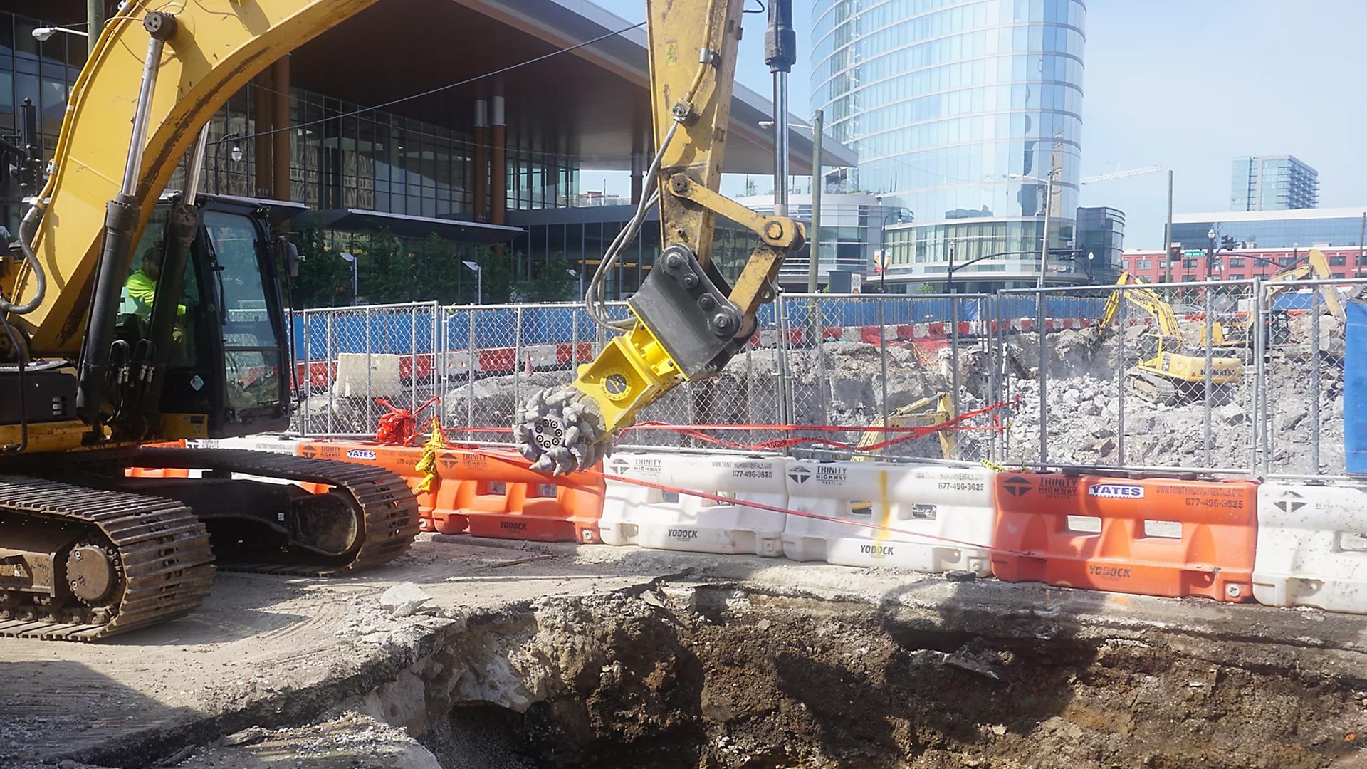 Yellow excavator with rotary cutter digs a trench at a construction site with barriers and distant buildings.